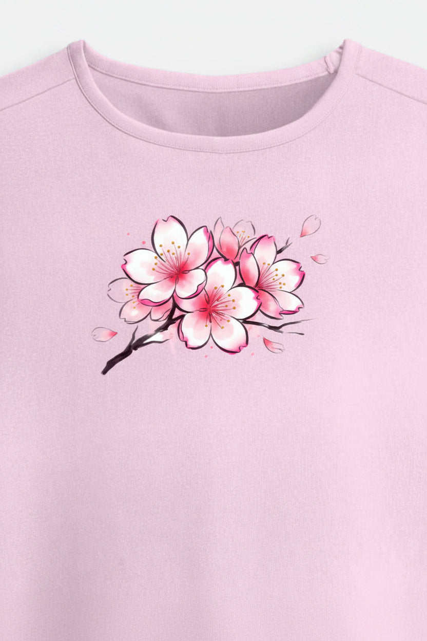 A light pink short-sleeve crop top t-shirt featuring a watercolor-style graphic of pink and white cherry blossoms on a branch across the chest, displayed on a clean off-white background.